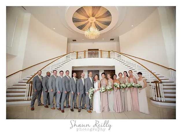 Wedding party posed in a grand white staircase foyer beneath a chandelier