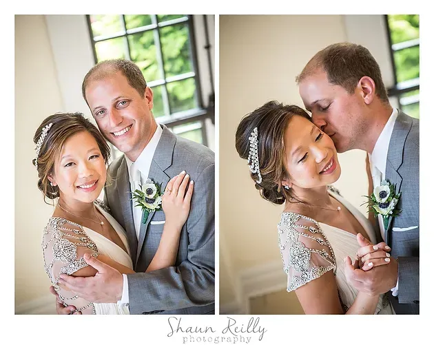 Smiling couple in wedding attire pose indoors, with the groom kissing the bride on the cheek.
