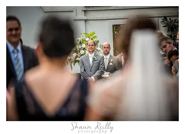 Groom and officiant at outdoor wedding, seen through blurred bride and guest in foreground