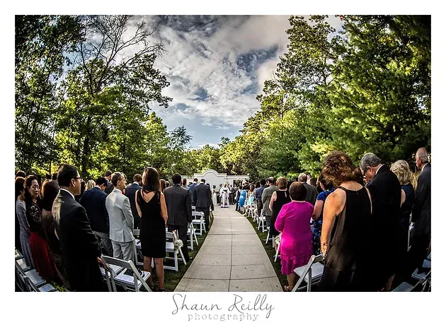 Guests line a central aisle outdoors under trees for a wedding ceremony, with one person in a bright pink dress.