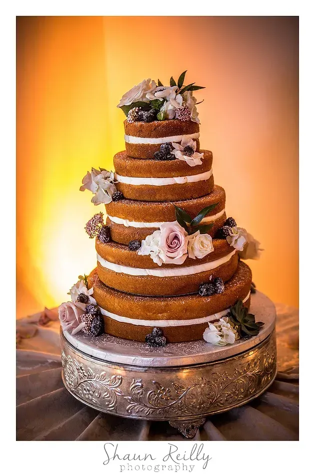 Tiered wedding cake with gold and purple floral accents on a decorative stand against a warm orange backdrop
