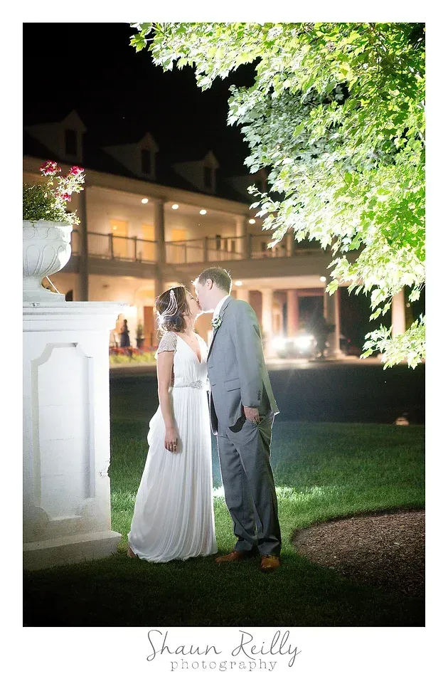 Bride and groom kiss under a lit tree outside a mansion at night.