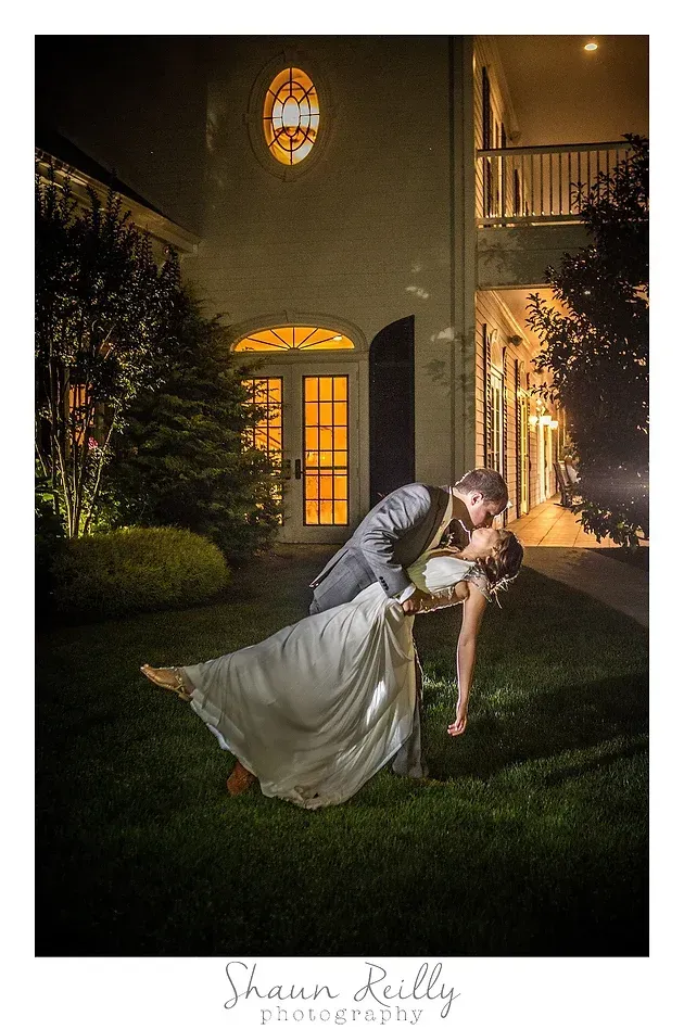 Wedding couple posing in a lit garden at night, bride in white gown being dipped by groom in tuxedo