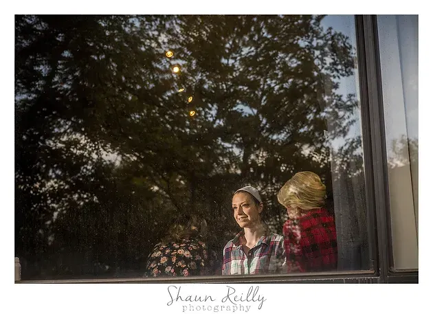 Two people sit by a window, reflected in the glass, with trees and warm lights outside.
