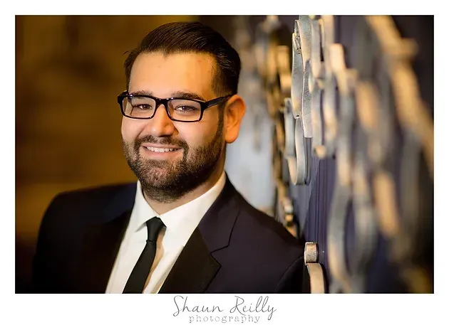 Smiling man in a dark suit and glasses, standing beside a blurred reflective wall.