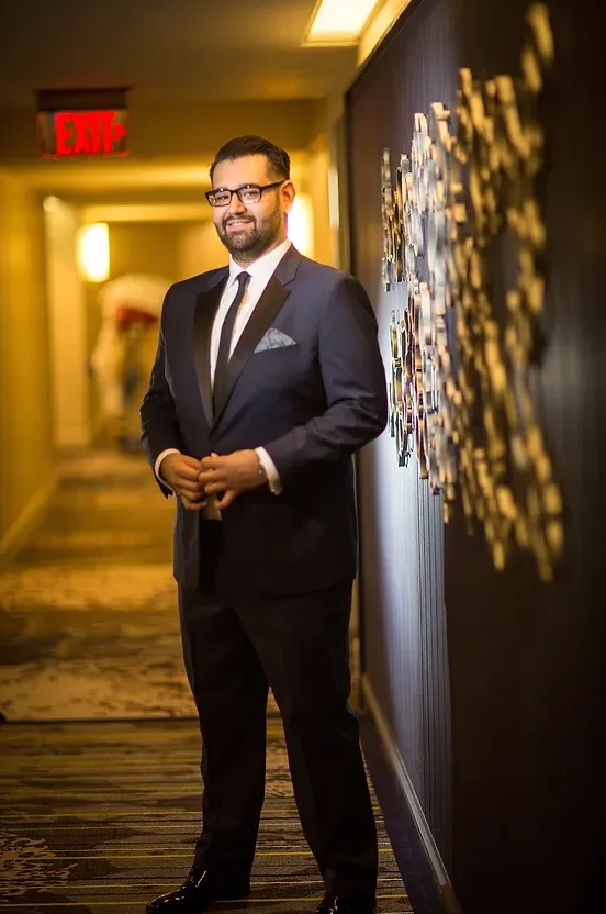 Man in a dark suit standing in a dim hallway beside a lit wall art display.