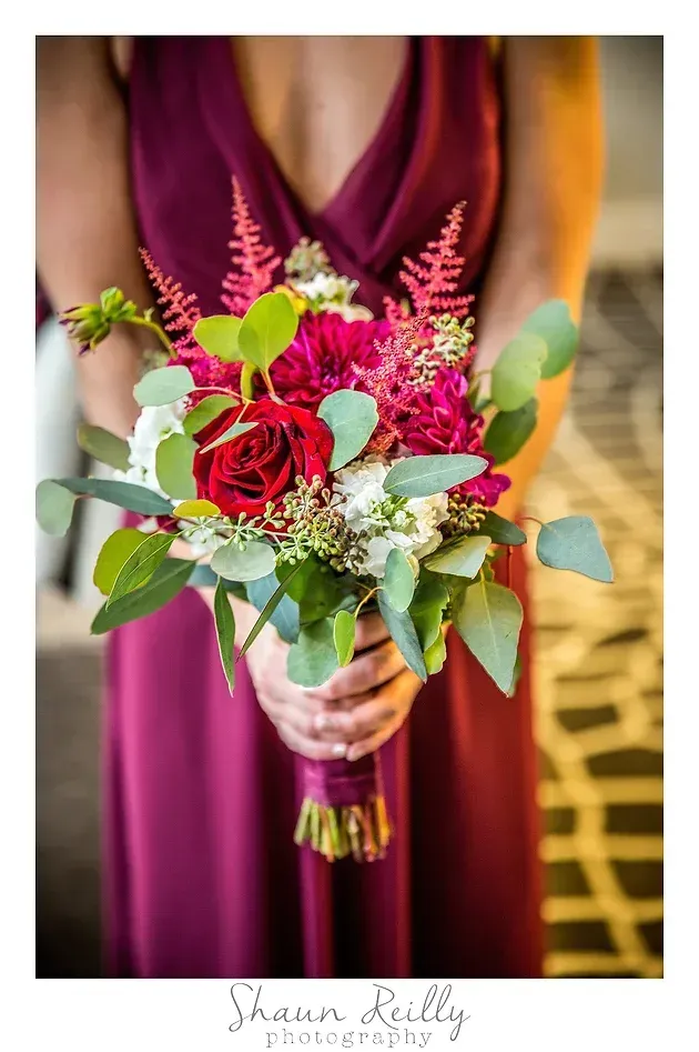 Bride holding a bouquet of red roses, pink blooms, and greenery against a burgundy dress