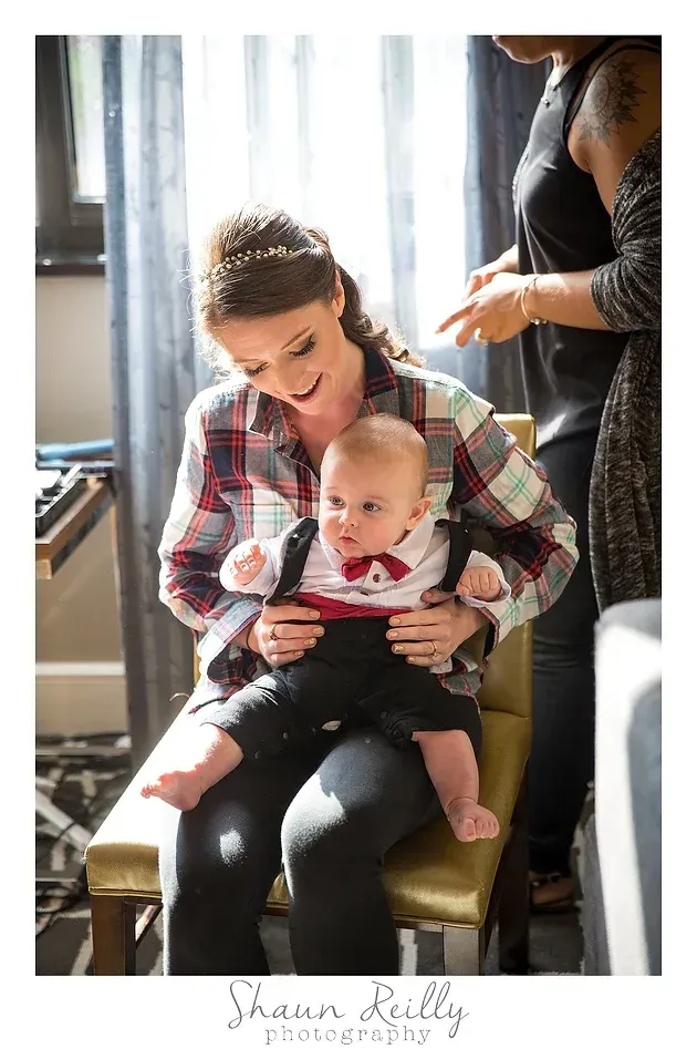 Woman holding a baby on a chair by a bright window, with another person standing nearby.