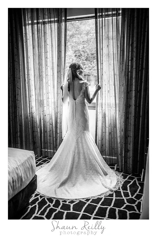 Bride in a white gown standing by a window with sheer curtains, holding the drapes and looking outside.