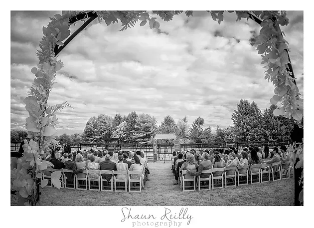 Outdoor wedding ceremony with guests seated under a floral arch, viewed from the back of the aisle
