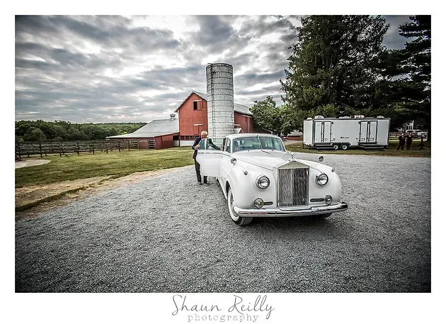 White vintage convertible on gravel beside a red barn and silo under a cloudy sky