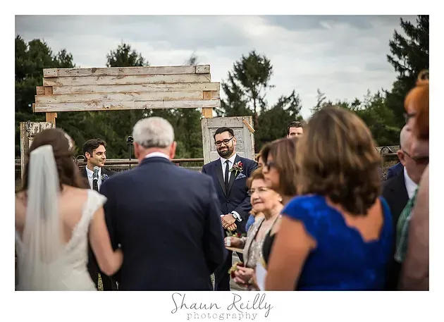 Bride and guests gathered outdoors at a rustic wedding ceremony, with a man speaking at center