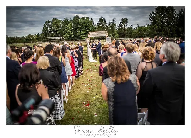 Outdoor wedding ceremony with guests lined along a grassy aisle under cloudy skies