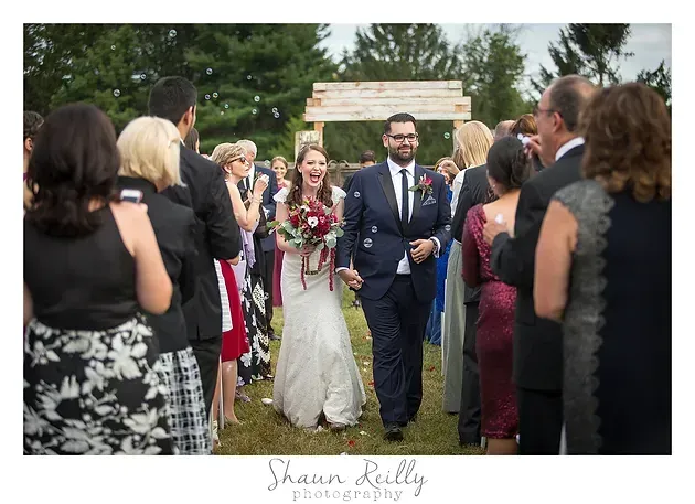 Bride and groom walking down an outdoor aisle, guests standing on both sides, with floral bouquets and greenery.