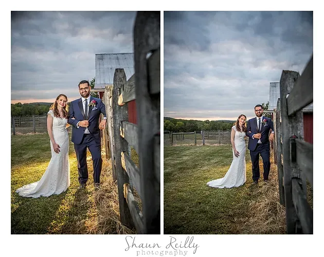Bride and groom pose outdoors by a stone wall under a cloudy sky, holding hands in wedding attire.