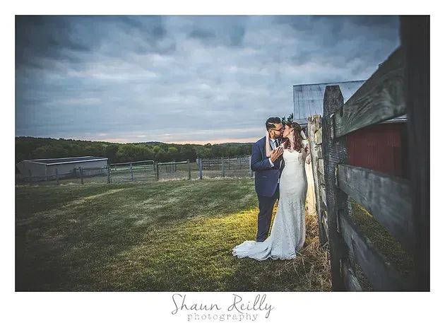 Bride and groom embracing outdoors by a wooden fence under a cloudy sky