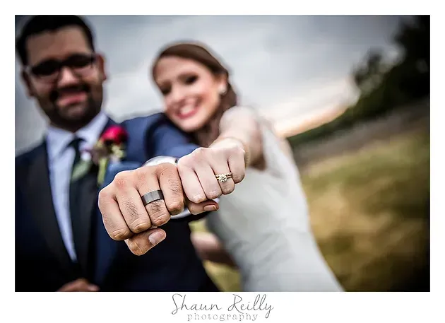 Newlyweds smiling outdoors, showing wedding rings close to the camera with a blurred field behind them