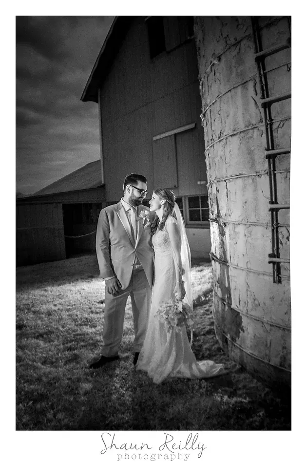 Wedding couple standing by a barn wall, the bride in a long gown and veil, in black and white.