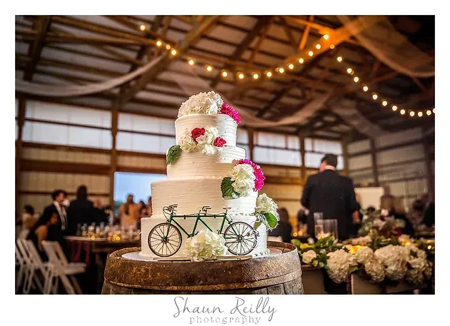 Three-tier white wedding cake with flowers and a bicycle topper in a rustic reception hall