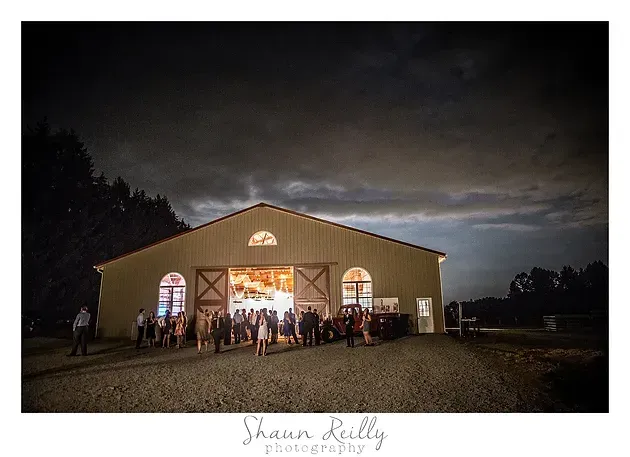Barn at dusk with people gathered outside under warm lights in a rural field.