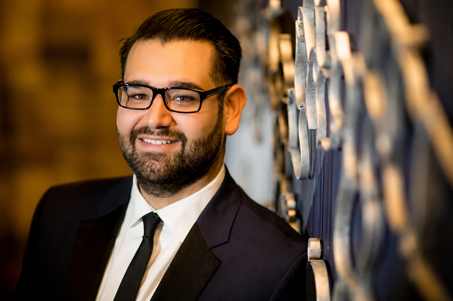 Smiling man in a dark suit and glasses standing beside a patterned wall