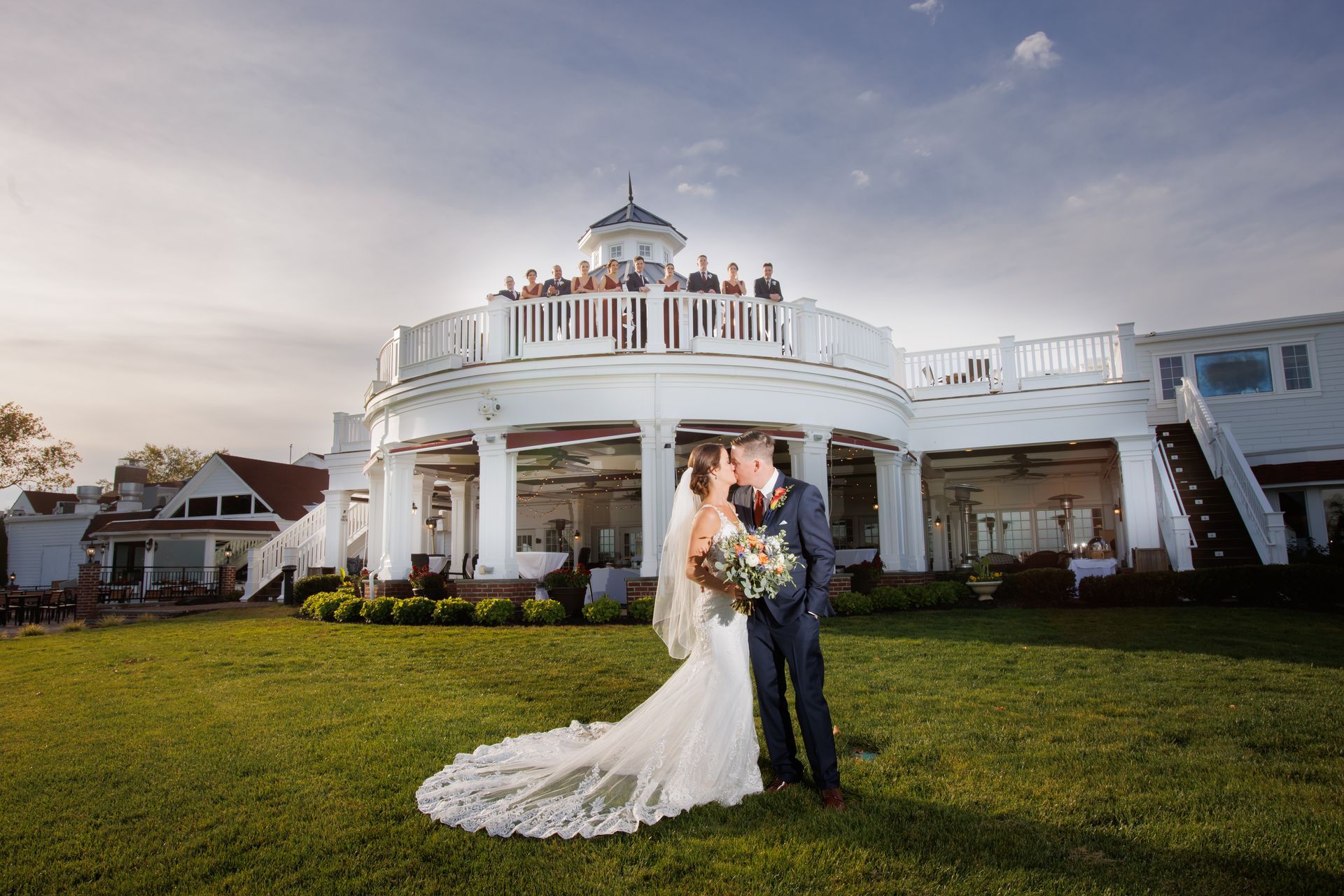 Bride and groom embrace on a lawn in front of a white mansion at sunset