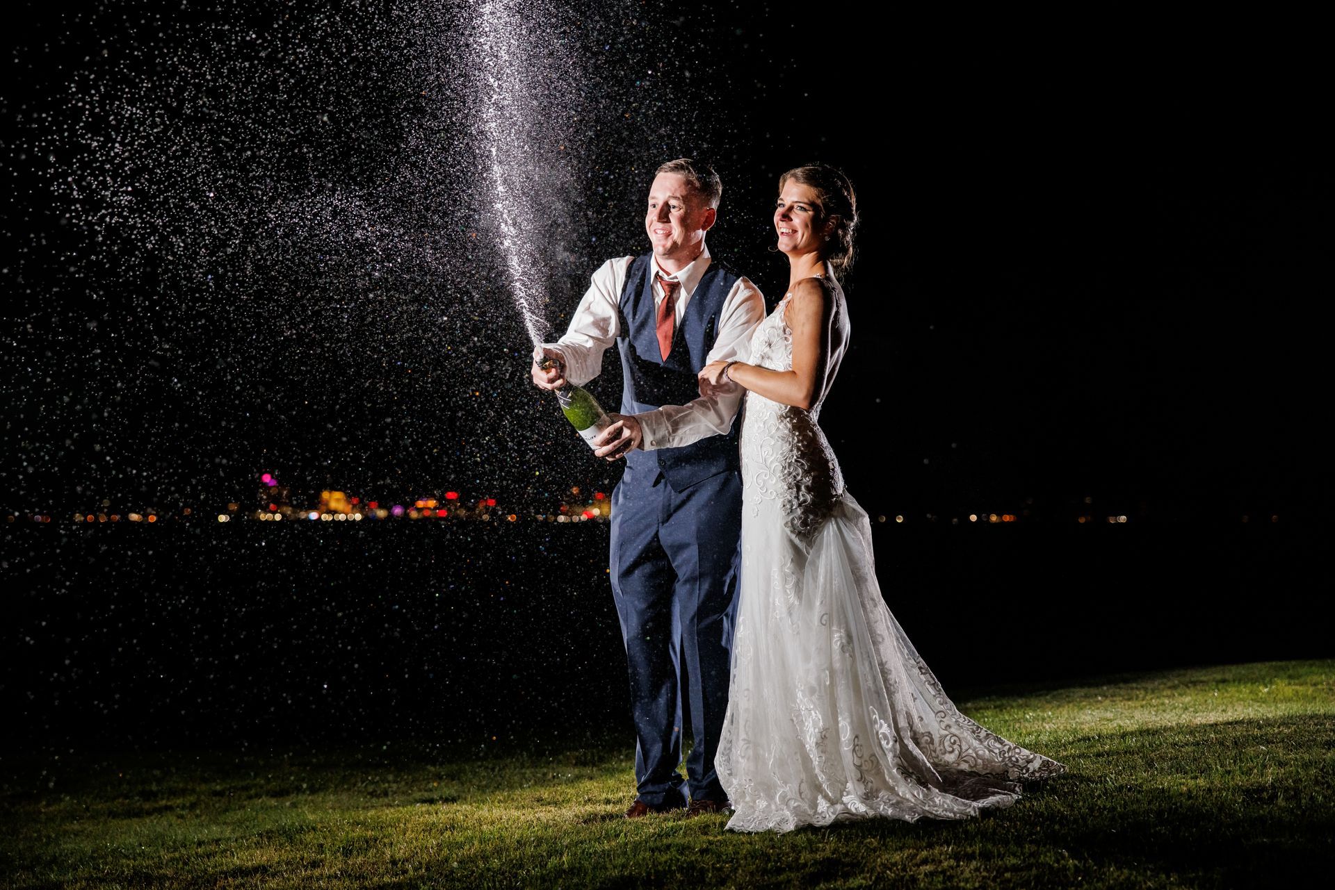 Newlyweds pop champagne on a lawn at night, with city lights glowing in the background.