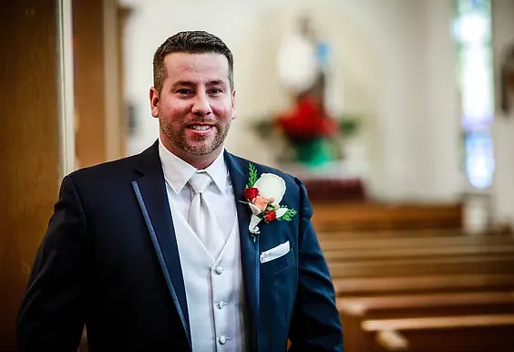 Man in a navy suit with a red boutonniere standing in a church aisle, smiling at the camera.