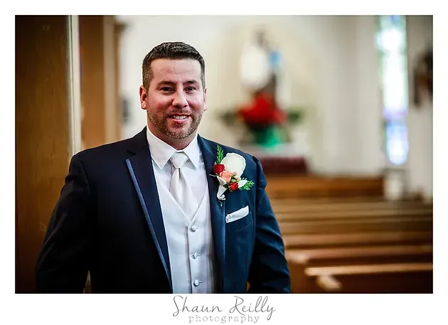 Smiling man in a navy suit and white vest standing in a church aisle with a boutonniere