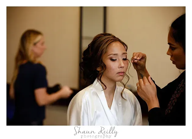Bride getting makeup applied while another woman stands in the background
