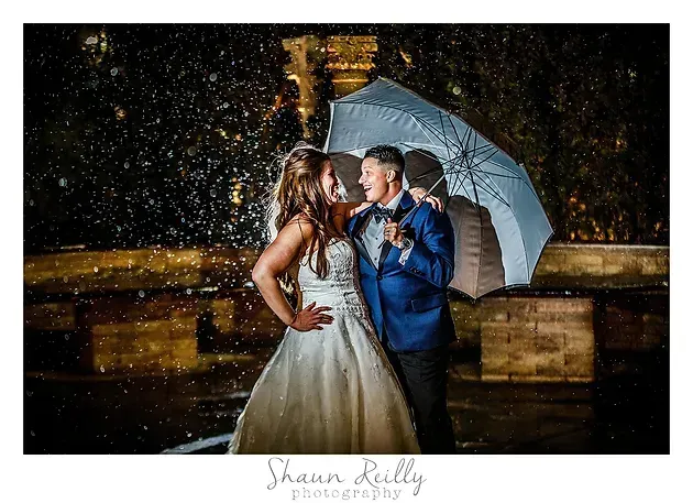 Bride and groom pose under a white umbrella in the rain at night, lit warmly outdoors.