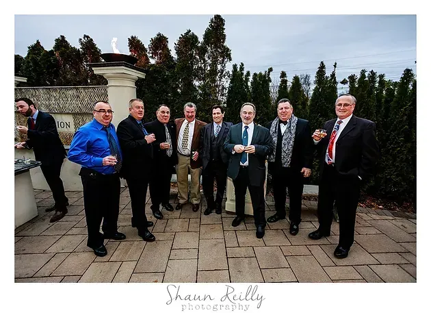 Group of men in suits posing outdoors on a stone patio, holding drinks and smiling.