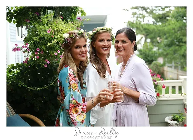 Three women smiling and holding drinks in a garden party setting, with flowers and greenery behind them