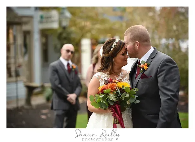 Bride and groom share a kiss outdoors, with a bouquet and a suited man blurred in the background.