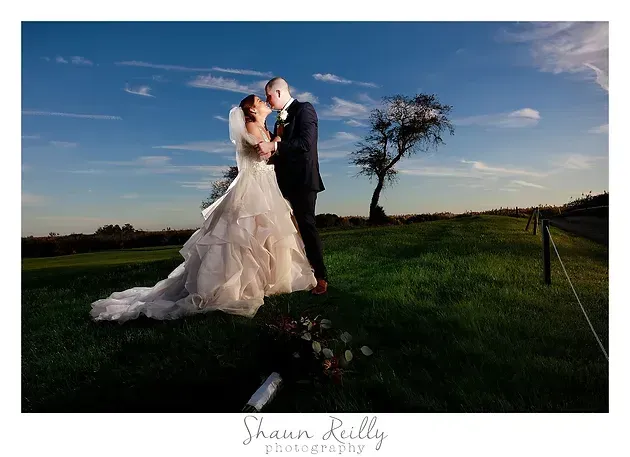 Bride and groom embrace on a grassy hill at sunset under a blue sky
