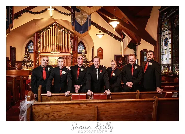 Seven men in black suits with red ties posing in a church pew under stained glass and wooden beams.