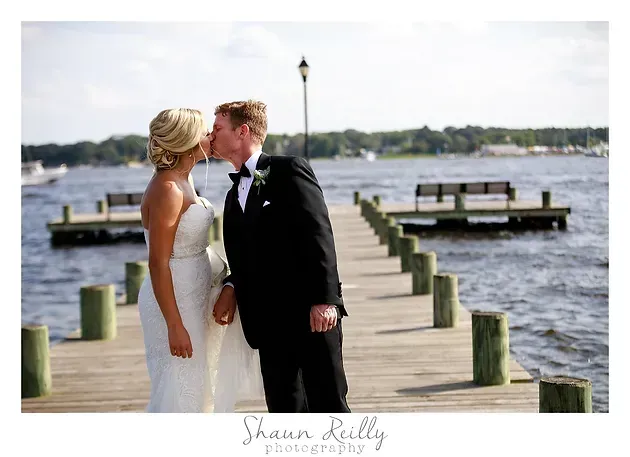 Bride and groom kissing on a dock by the water, with boats and a lamppost in the background