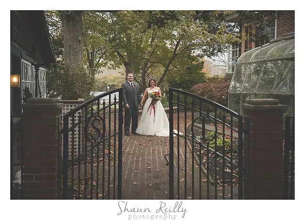 Bride and groom standing on a brick path by wrought-iron gates, holding hands in a garden setting