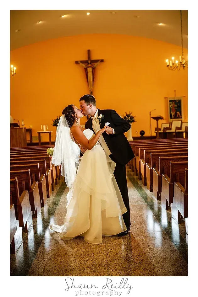 Bride and groom kissing in a church aisle beneath a cross, with warm lighting and empty pews