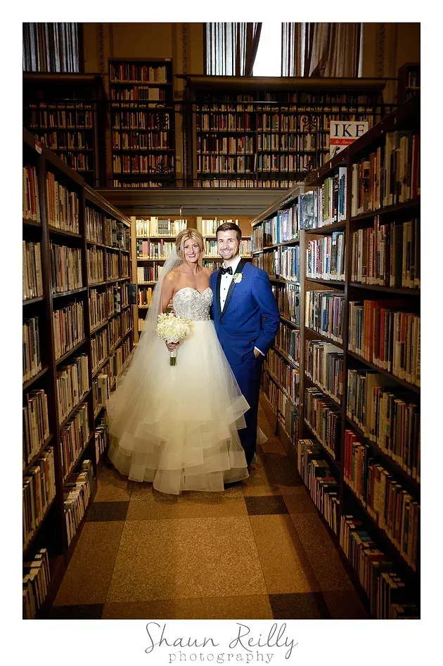 Bride and groom standing in a narrow library aisle, surrounded by tall bookshelves.