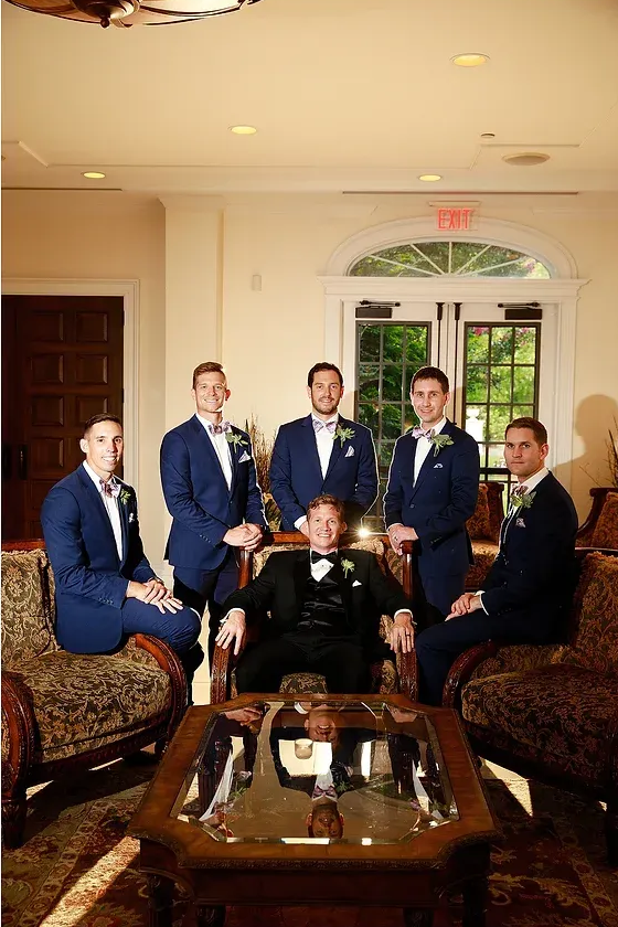 Six men in suits posing in a formal room with a coffee table and ornate chairs.