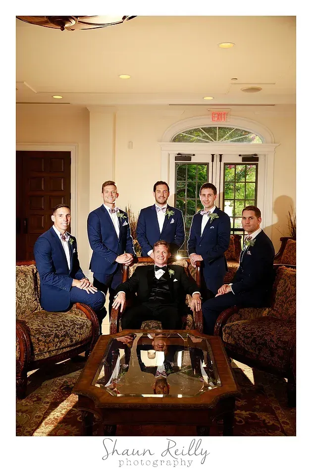 Five men in navy suits pose in a formal living room around a coffee table.