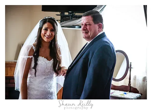 Bride in white dress and veil standing beside a smiling man in a blue suit indoors.