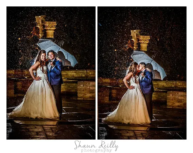 Bride and groom pose under an umbrella in the rain at a lit outdoor fountain at night