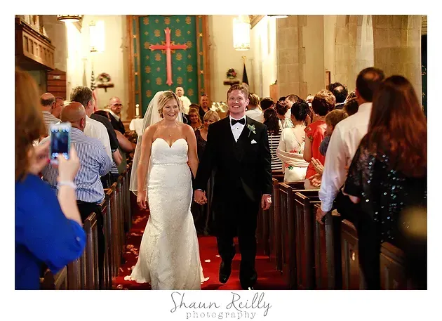 Bride and groom walk down the aisle in a church, with guests lining the pews and a green altar backdrop.