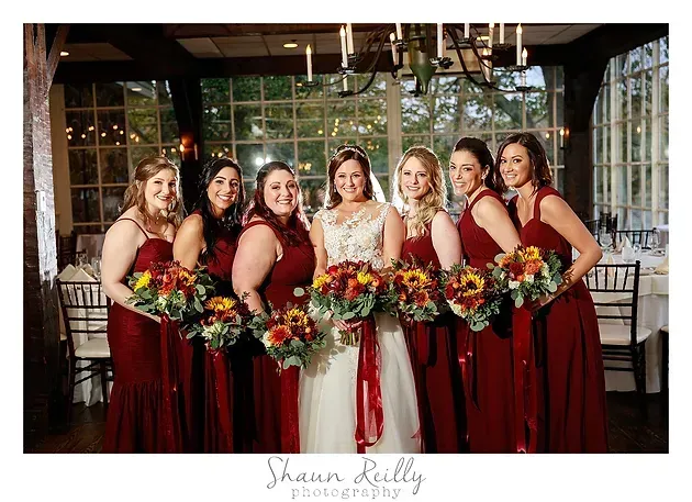 Seven bridesmaids in burgundy dresses holding bouquets, standing indoors in front of large windows.