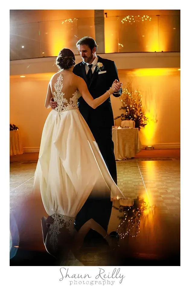 Bride and groom slow dancing in a warmly lit hall, reflected on the polished floor