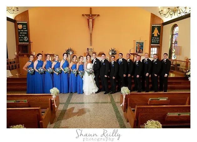 Wedding party posed in a church aisle with bride and groom, bridesmaids in blue, and groomsmen in black