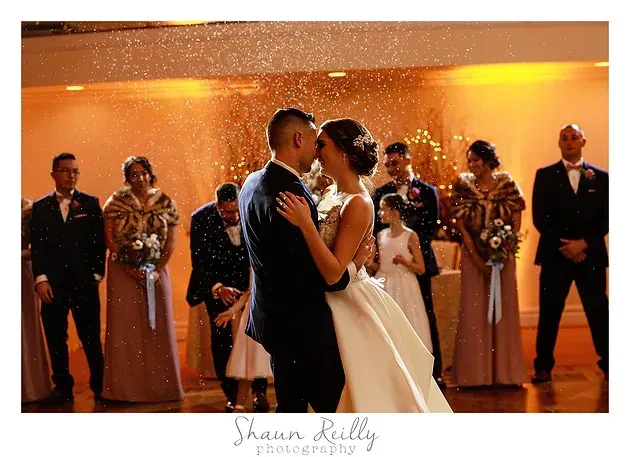 Bride and groom share a dance at a warm-lit reception, with wedding party standing behind them.
