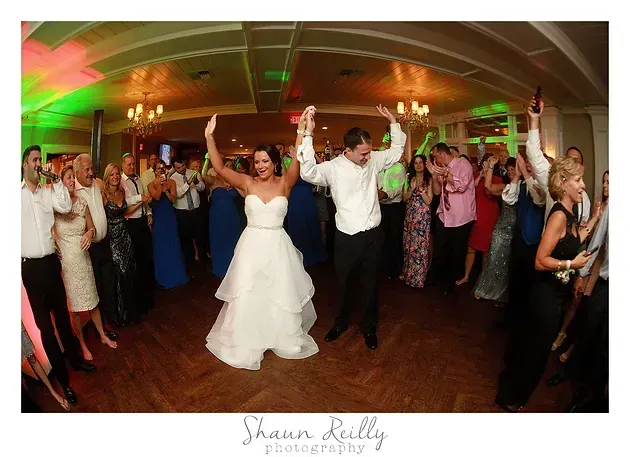 Wedding couple dancing with raised arms at a lively reception, guests cheering around them